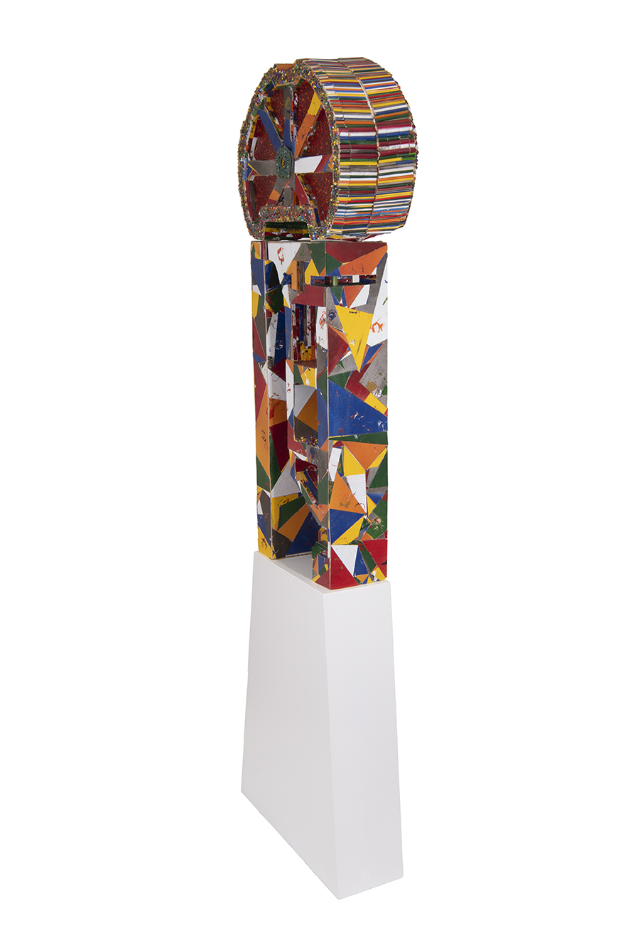 A freestanding sculpture featuring a large circular wheel at the top, decorated with multicolored radial segments and bordered by evenly spaced vertical strips. The wheel sits atop a tall rectangular column covered in a patchwork of brightly painted geometric shapes, mounted on a plain white pedestal.