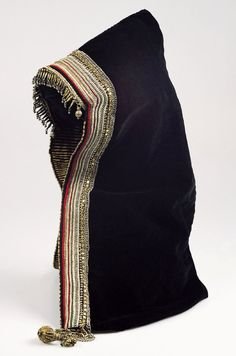 Studio photo of a black hooded garment or veil draped upright, trimmed with gold fringe and red-and-gold embroidered bands along the edge against a white background.