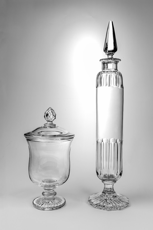 Two empty glass jars with lids are displayed against a plain light background. The jar on the left is short and wide with a rounded lid, while the jar on the right is tall and narrow with a pointed lid.