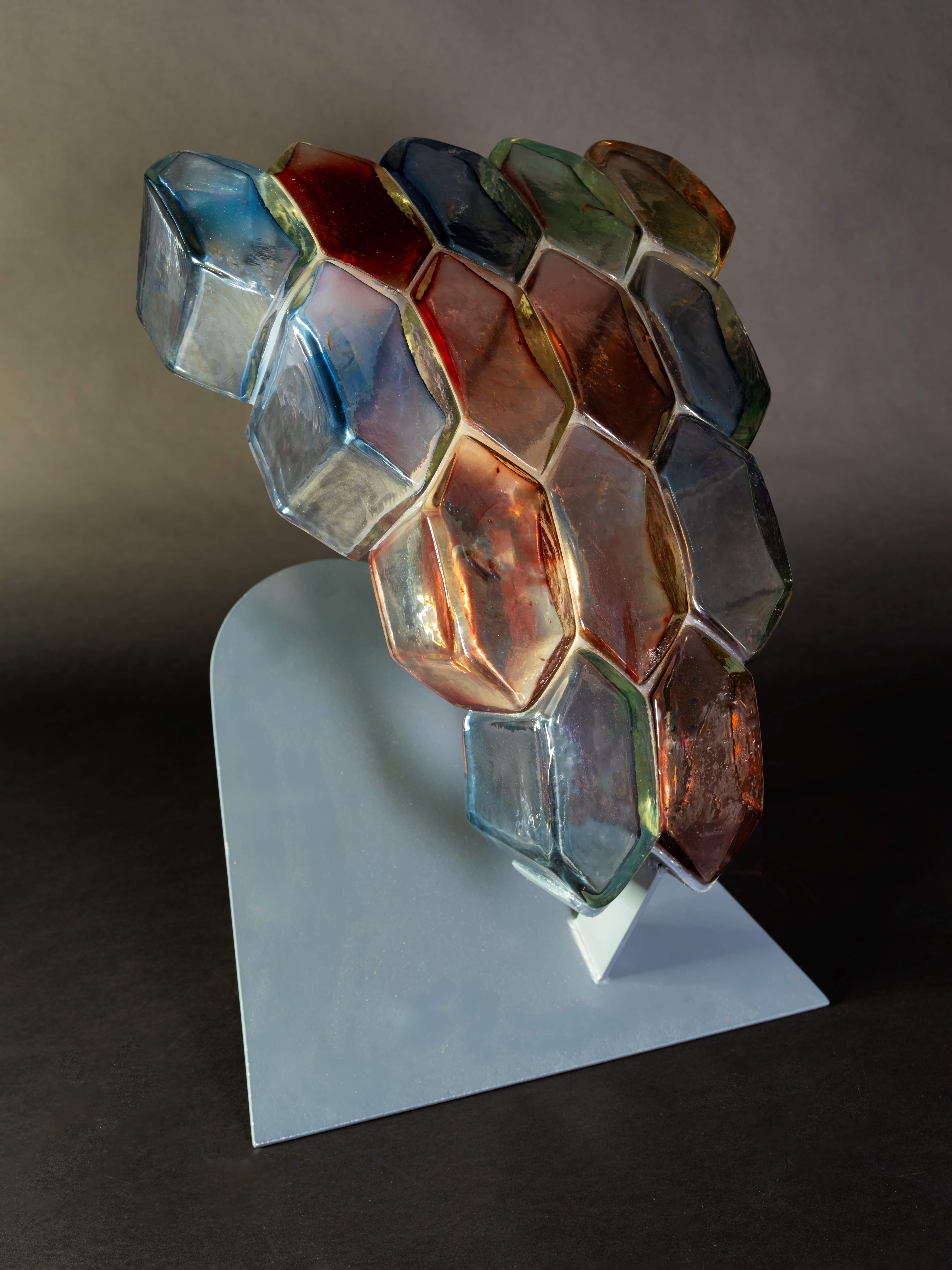 Abstract sculpture featuring a cluster of hexagonal glass shapes in a honeycomb pattern. The glass elements are tinted in various colors including blue, red, green, and amber, and are arranged closely together. The structure is supported by a light gray, angled metal base, which contrasts with the dark background. Light reflects off the colored glass, highlighting the geometric pattern and vibrant hues.