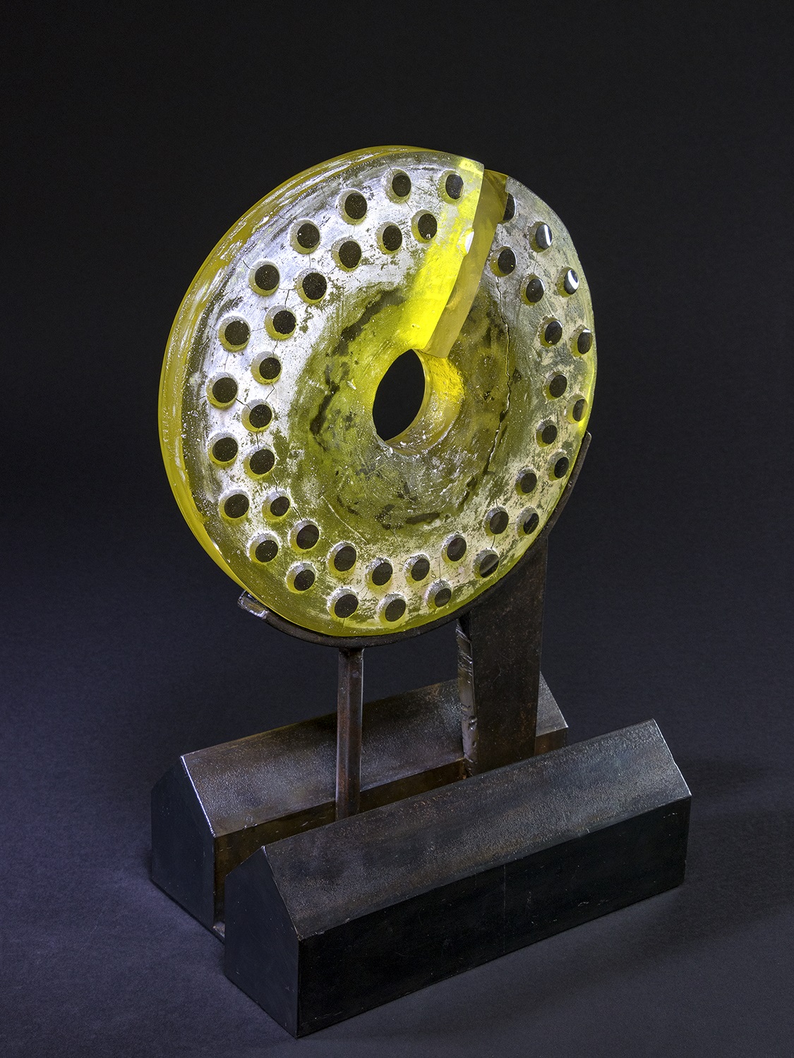 Circular sculpture made of yellow-tinted translucent material with a textured surface, decorated by rows of circular holes throughout. The disc has a central opening and a single wedge-shaped cut running from the edge to the center. It is mounted upright on a dark, rectangular metal base using two vertical metal supports. The background is plain black, emphasizing the bright and intricate details of the sculpture.