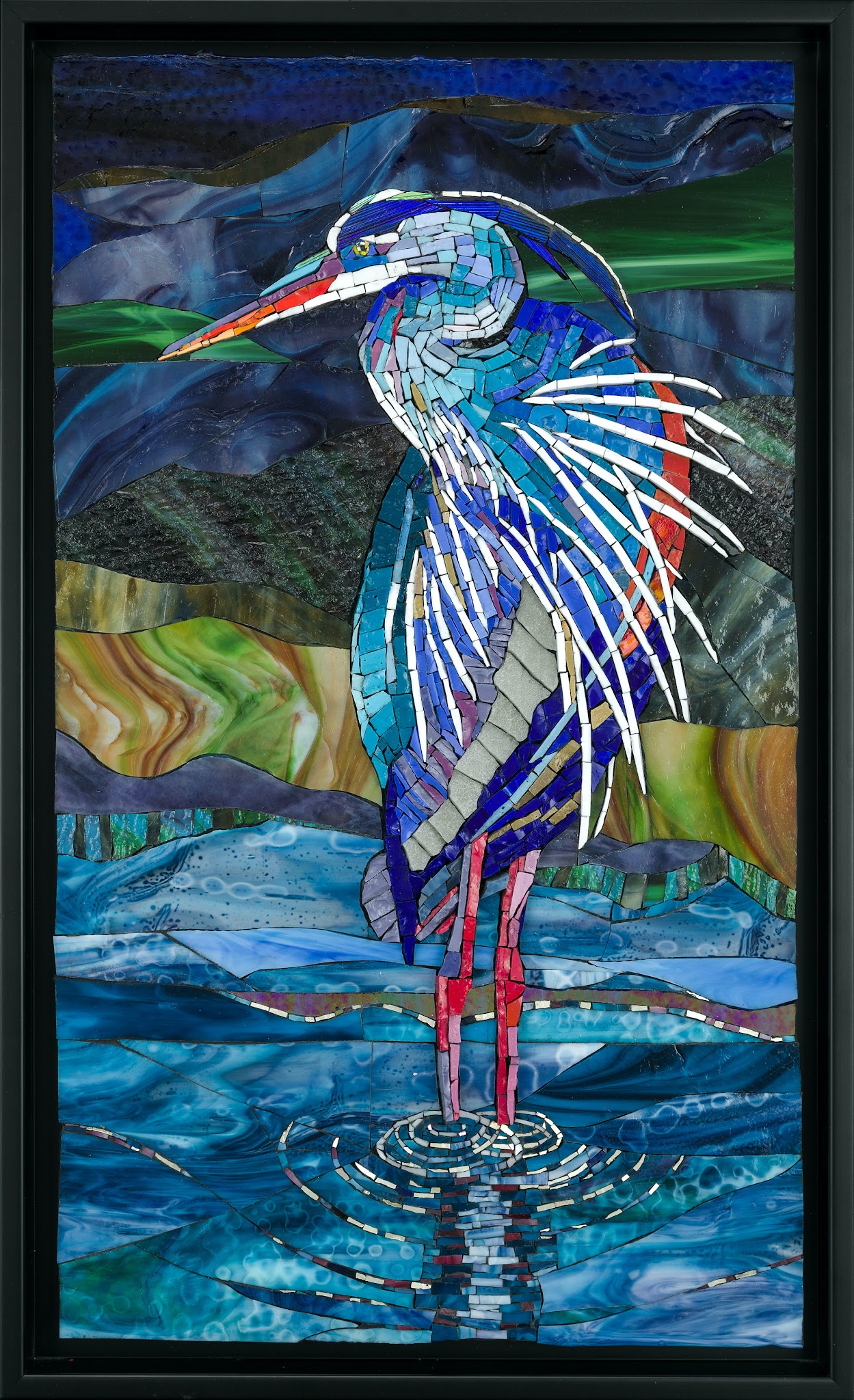 ramed glass mosaic of a blue heron standing in shallow water, with a long orange beak, layered blue-and-white feathers, and pink legs creating ripples; abstract bands of dark sky and green-and-brown landscape fill the background.