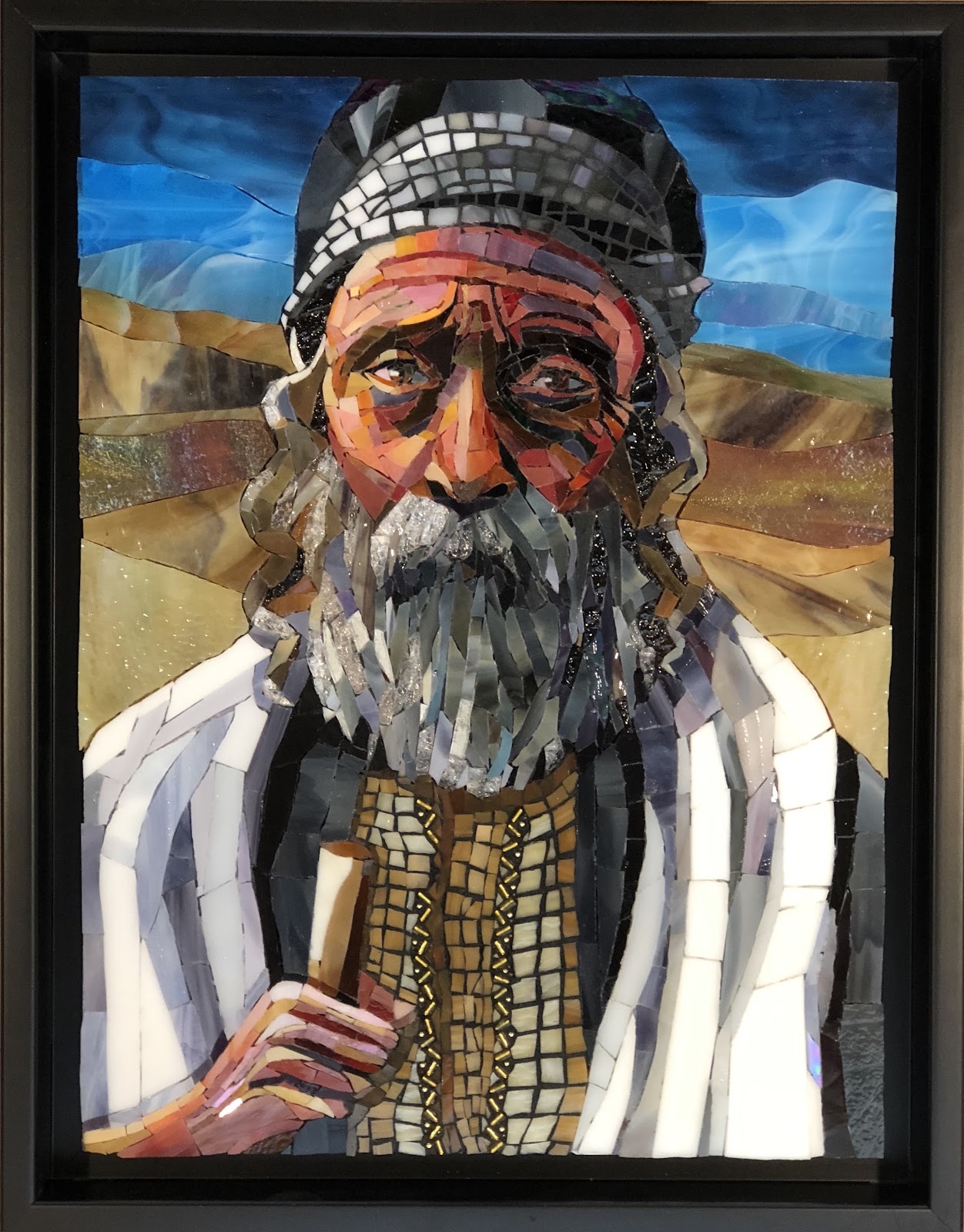 Framed glass mosaic portrait of a person with the face obscured by a rectangular blur. The figure wears a patterned head covering and a white-and-gray draped garment over a gold, grid-like vest, holding one hand near the chest. Behind them, layered bands of tan and brown suggest hills beneath a bright blue sky.