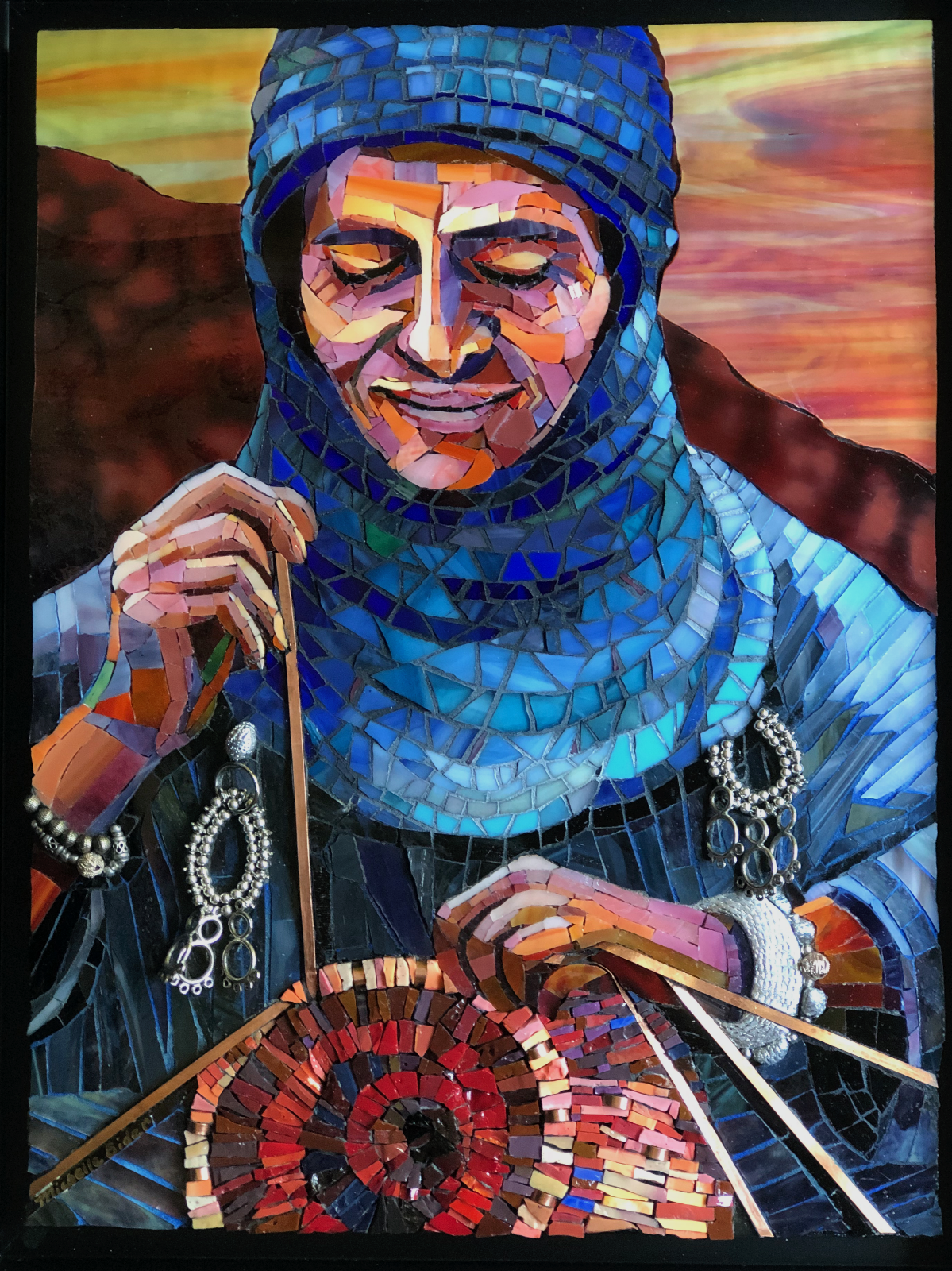 Glass mosaic portrait of a person with the face obscured by a rectangular blur, wearing a blue, tile-like headscarf and layered blue clothing. One hand holds a thin rod or reed, while the other rests near a red-and-brown coiled basket in the foreground, doing basket weaving. Silver jewelry hangs at the wrists, and dark hills sit behind the figure under a streaked, multicolored sky.
