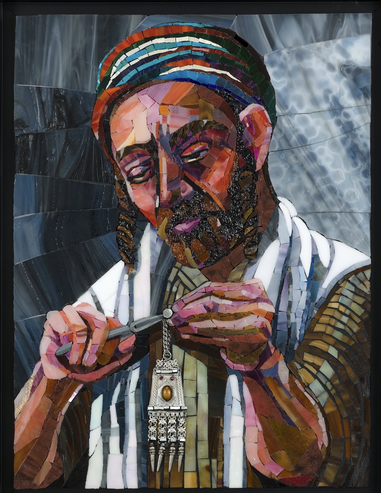 Framed glass mosaic portrait of a jeweler with the face obscured by a rectangular blur, wearing a multicolored striped cap and a white-and-blue striped wrap. He holds a small tool and metal piece at chest height, with a detailed silver pendant featuring an amber-colored stone hanging below. The background is composed of swirling gray-blue glass pieces.