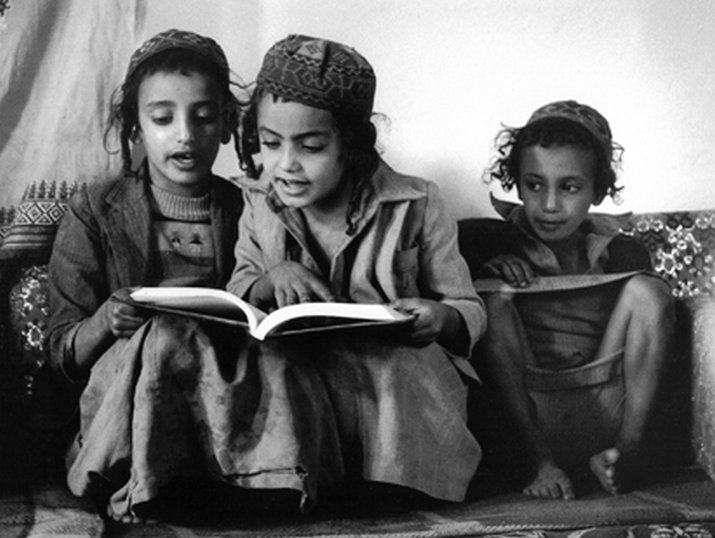 Black-and-white photo of three children sitting on cushions indoors, wearing patterned caps and layered clothing; the middle child holds an open book while the others look on, and their faces are obscured.