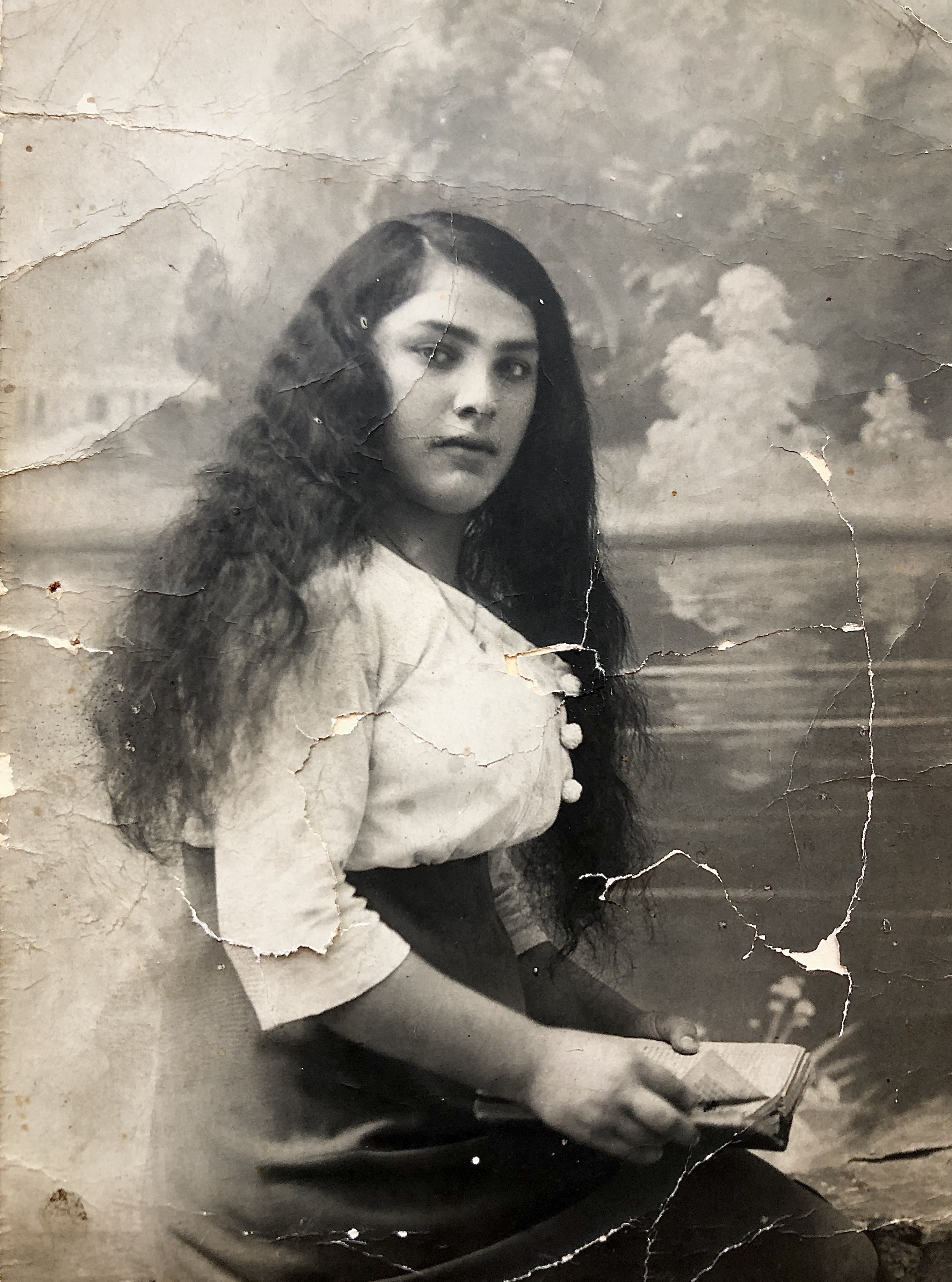 Worn black-and-white vintage photograph of a seated person with long dark wavy hair holding a small book or notebook, posed in front of a painted landscape backdrop; the photo is creased and torn, and the face is obscured.