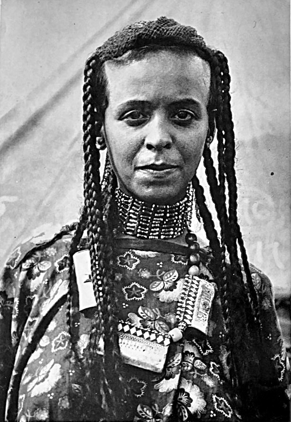 Black-and-white portrait of a person with long braided hair, wearing a beaded face covering and a patterned garment with decorative necklaces and pendants; the face is obscured.