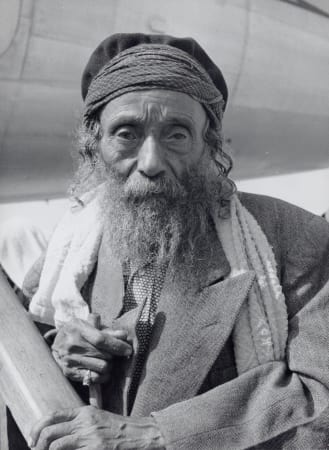 Black-and-white photo of a bearded person wearing a cap and layered clothing with a scarf draped over one shoulder, leaning forward and holding an object near their chest; the face is obscured.
