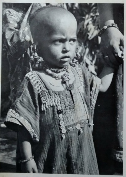 Black-and-white photo of a small child wearing a patterned tunic and multiple necklaces and bracelets, standing outdoors while holding an adult’s hand; the child’s face is obscured.