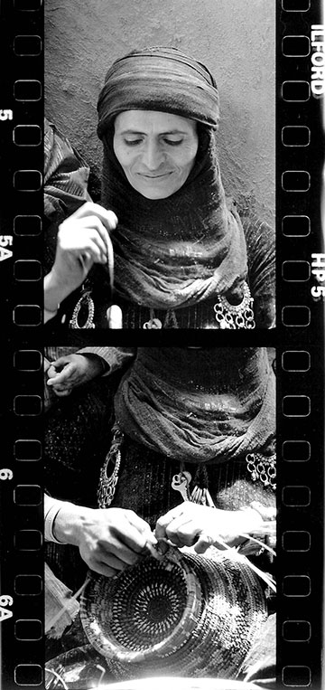 Black-and-white film-strip image of a seated person with their head and face wrapped in a scarf, wearing patterned clothing with coin-like decorations and holding a woven basket; the face is obscured.