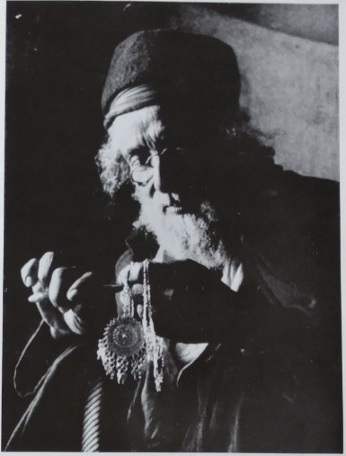 Framed glass mosaic portrait of a jeweler with the face obscured by a rectangular blur, wearing a multicolored striped cap and a white-and-blue striped wrap. He holds a small tool and metal piece at chest height, with a detailed silver pendant featuring an amber-colored stone hanging below. The background is composed of swirling gray-blue glass pieces.