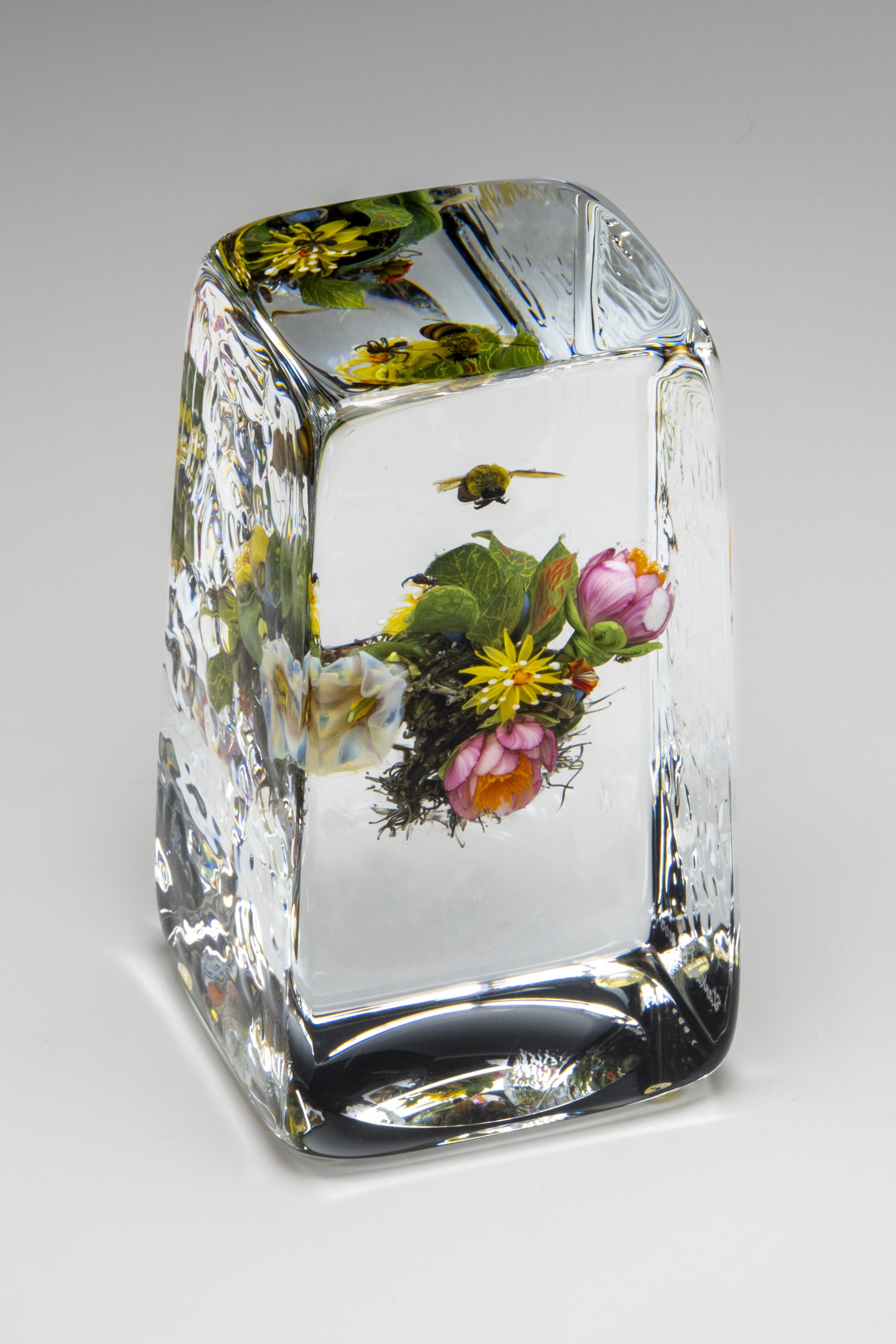  A clear glass paperweight featuring a detailed, realistic depiction of colorful flowers and a bee suspended inside. The paperweight has a rectangular shape with rounded edges.