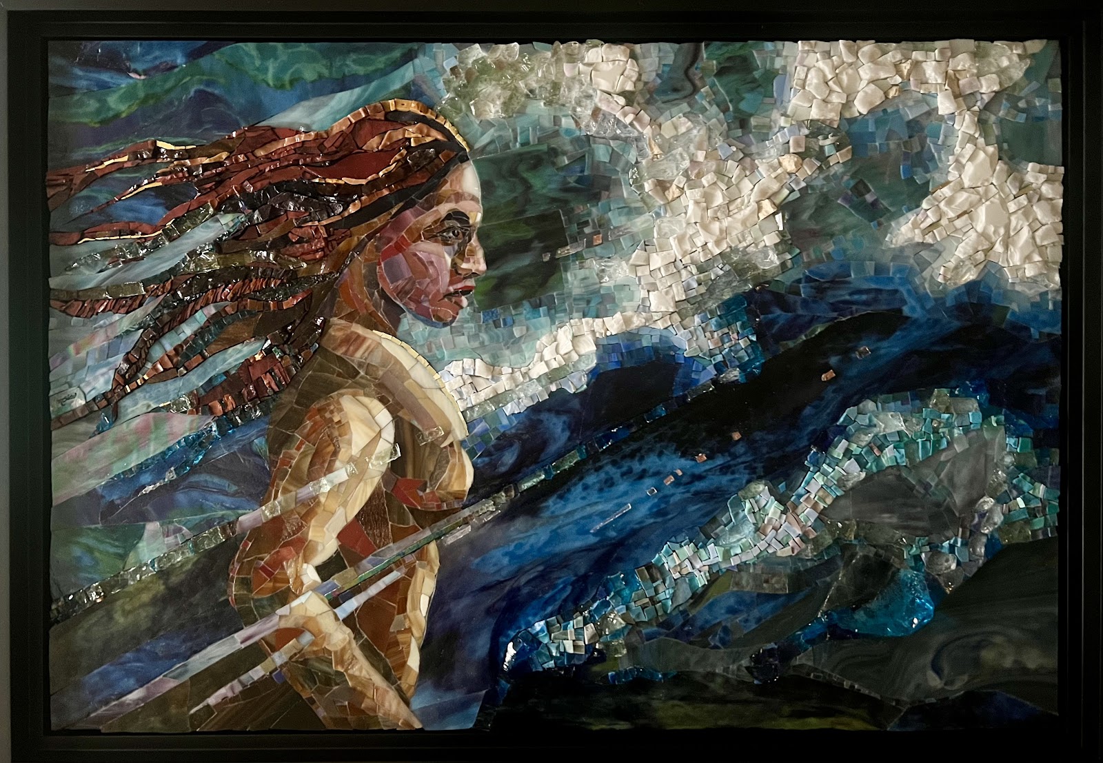Framed mosaic artwork of a swimmer in rough water, shown from the side with long flowing hair and an arm reaching forward. The scene is built from layered blue and green glass pieces, with bright white, foamy waves breaking across the upper right.