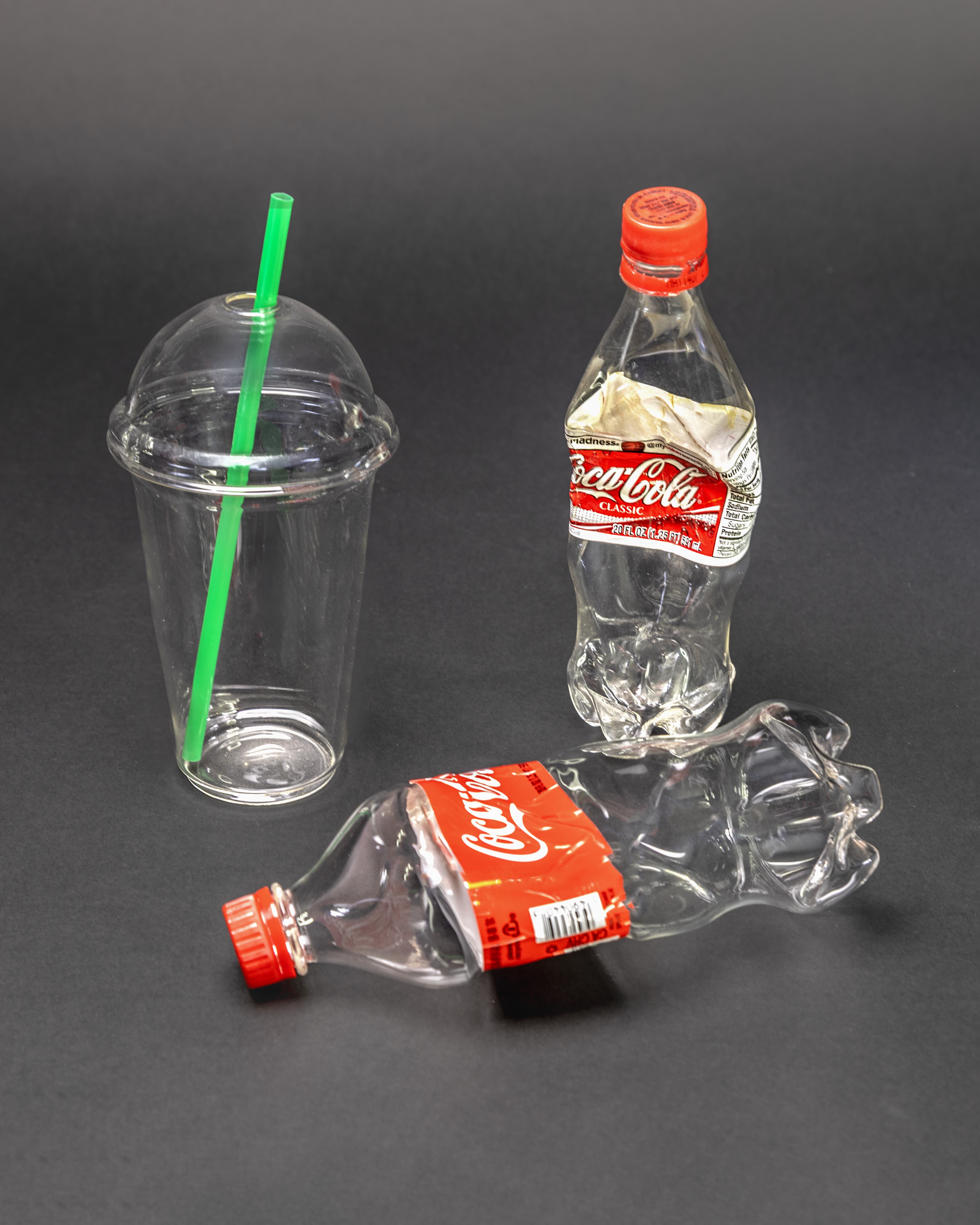 Three empty plastic containers on a dark background: a clear plastic cup with a green straw and domed lid, and two crumpled Coca-Cola bottles with red caps and labels—one standing upright and one lying on its side.