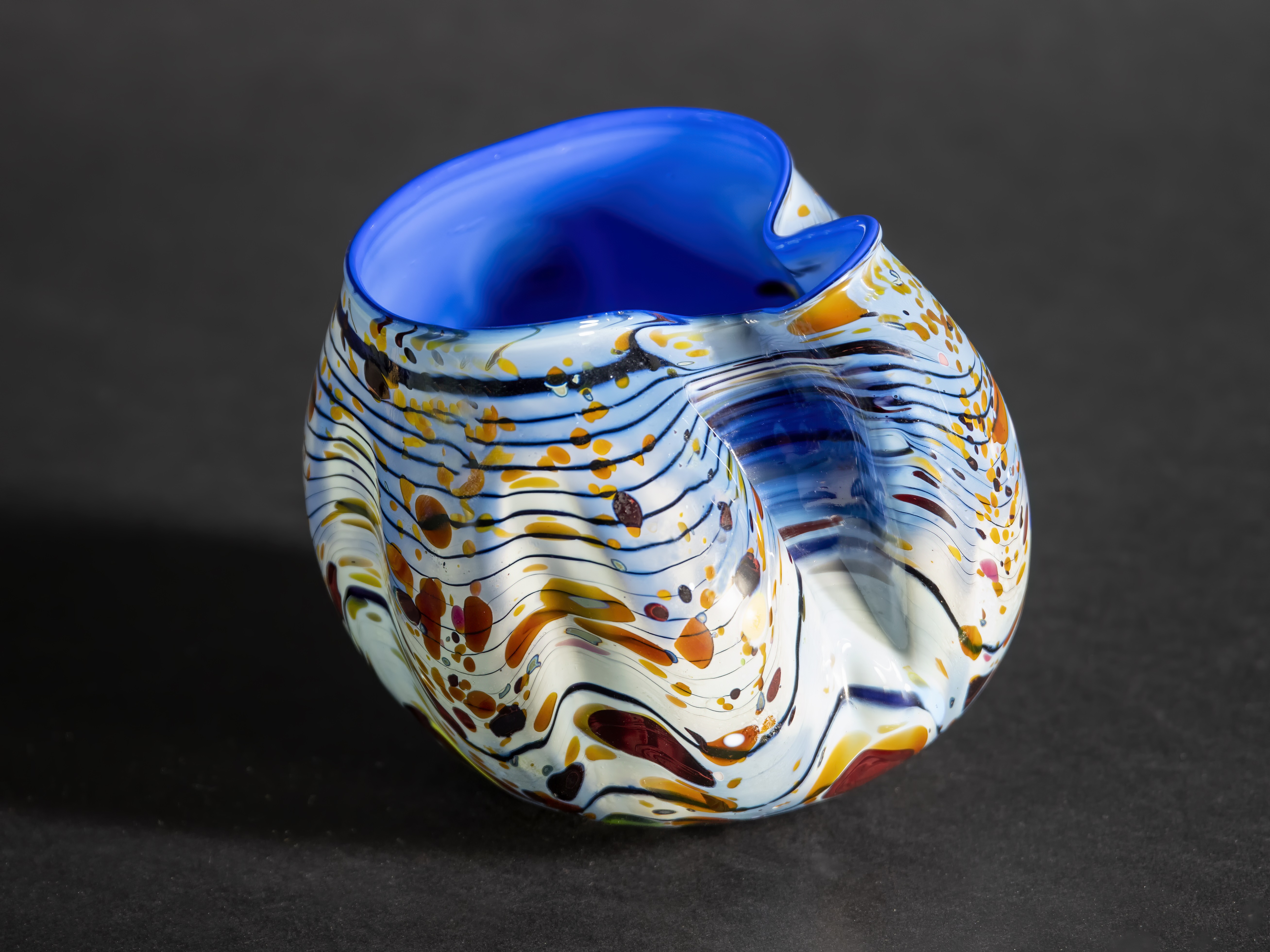 An artistic, brightly colored glass vessel with a vivid blue interior and an irregular, organic shape. The exterior features swirling lines and speckles in blue, orange, red, and black, creating a dynamic, abstract pattern.