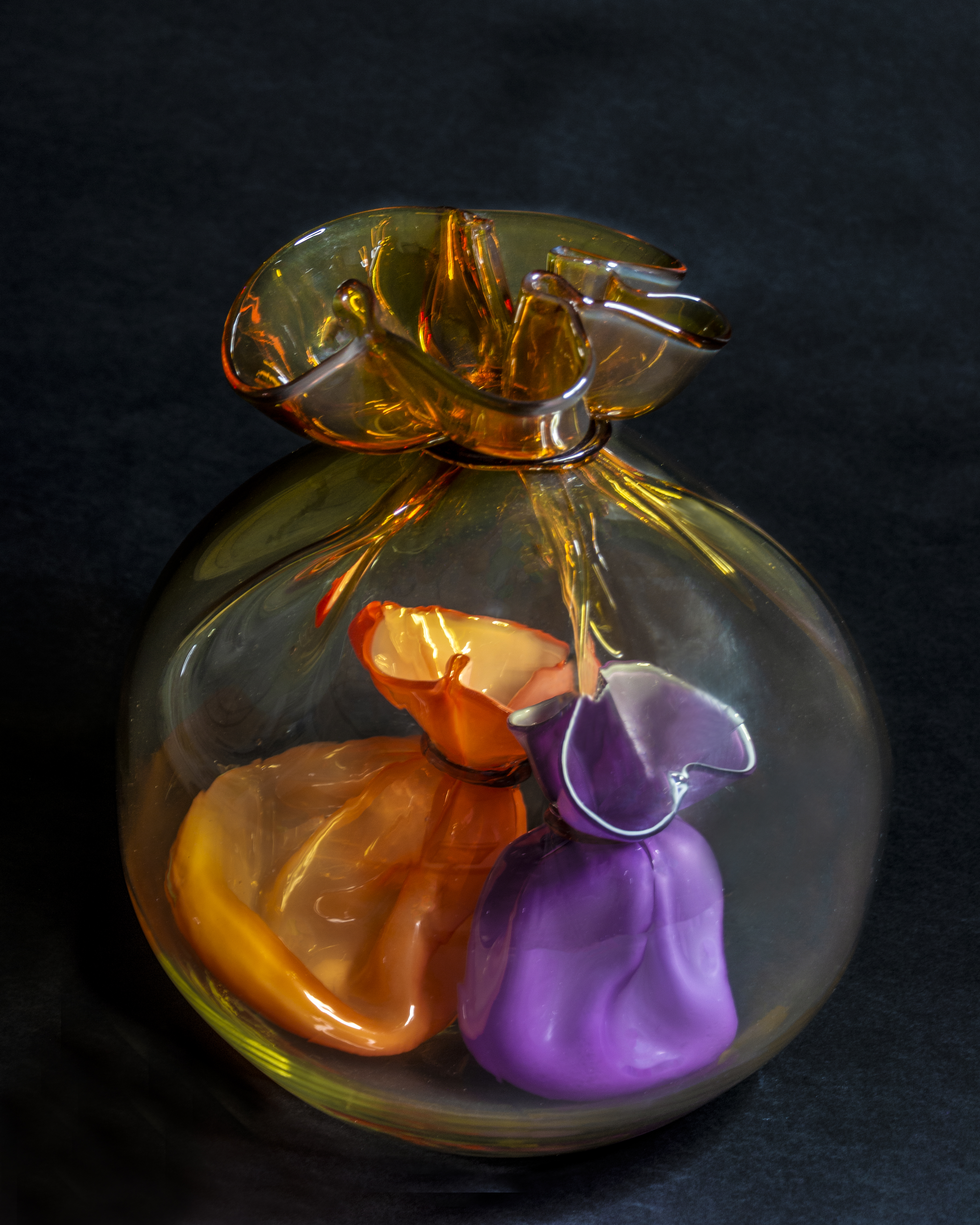 A glass sculpture shaped like a translucent bag tied at the top, containing two smaller bag-like shapes inside—one orange and one purple. The sculpture rests on a dark background.